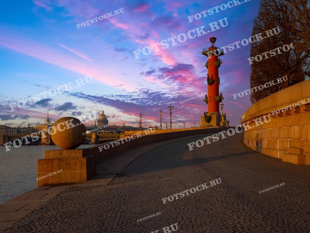 На фотографии изображена Ростральная колонна на Стрелке Васильевского острова в Санкт-Петербурге. Закатное небо раскрашивает облака в розовые и фиолетовые оттенки, создавая волшебную атмосферу. На заднем плане видны Адмиралтейство и величественный купол Исаакиевского собора, дополняющие живописный городской пейзаж.